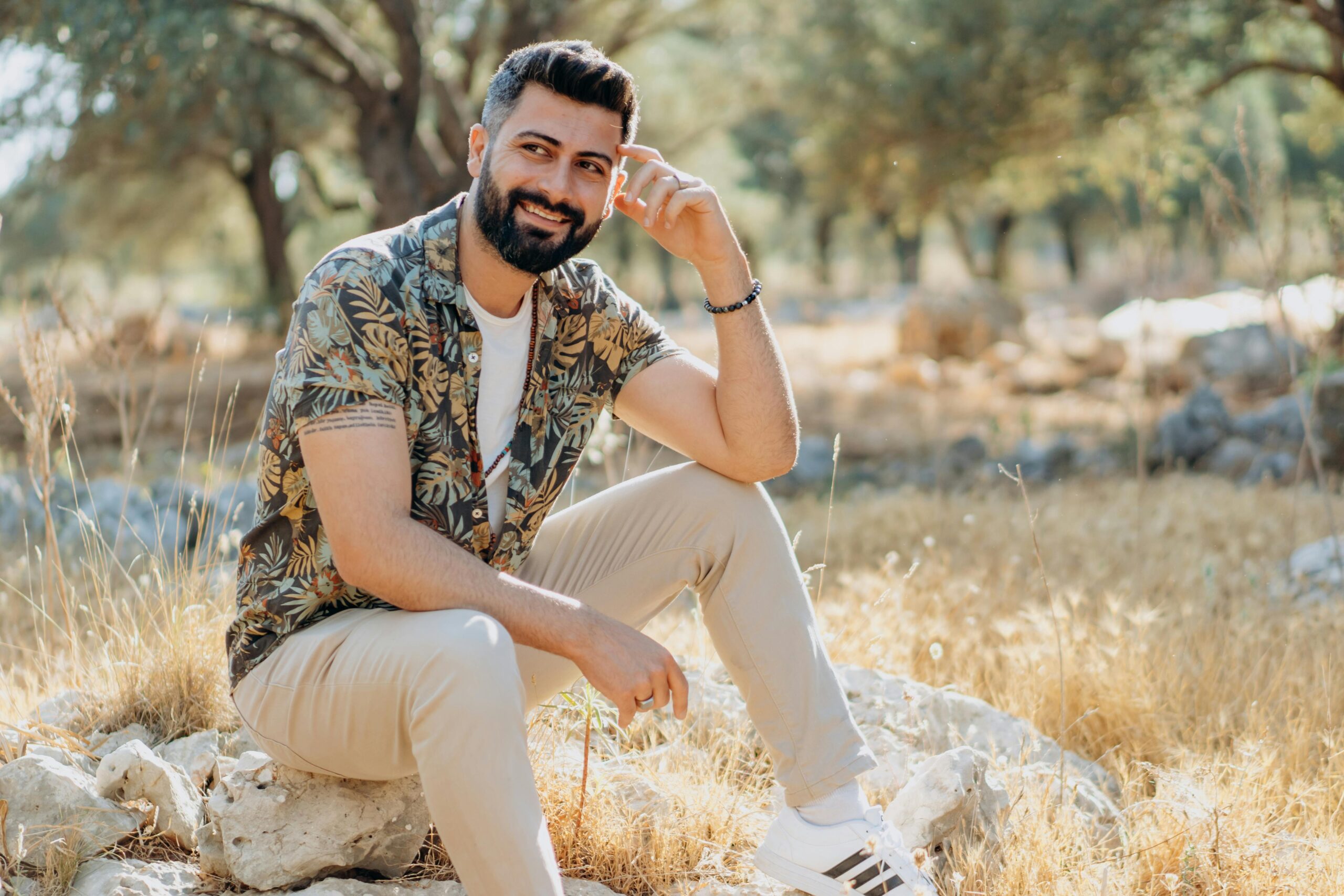 Home A fashionable man with a beard smiling while sitting outdoors in sunny nature.